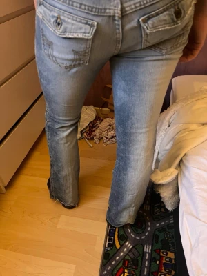Ljusblå bootcut jeans med fickdetaljer - Säljer ett par ljusblå bootcut jeans med snygga fickor och dekorativa sömmar på bakfickorna. Jeansen har en klassisk femficksdesign och är tillverkade i denim med lätt slitning för en cool look. Från Tommy hilfiger. Fransat sig nere vid ena benet men går att laga!! 