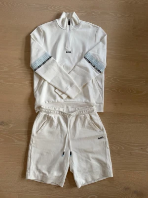 Vitt set från BOSS med tryck - Snyggt vitt set från BOSS med långärmad tröja med halv dragkedja och matchande shorts. Tröjan har BOSS-tryck på bröstet och armarna, samt detaljer i grått och svart. Shortsen har dragsko i midjan och BOSS-tryck på benet och bakfickan. Perfekt för en avslappnad look.