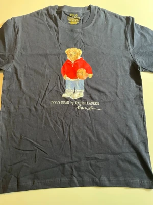 Svart Polo Bear t-shirt från Ralph Lauren - Snygg svart t-shirt från Ralph Lauren med ikoniska Polo Bear-trycket framtill. Björnen har på sig en röd hoodie och håller en basketboll. Klassisk rund hals och korta ärmar. Perfekt för dig som gillar streetstyle och vill ha något unikt i garderoben.