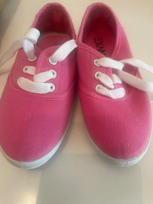 Rosa låga tygsneakers med vita snören - Snygga rosa sneakers i tyg med vita snören och vit sula. Klassisk låg modell som passar perfekt till jeans eller shorts. Skorna har rund tå och enkel design som gör dem lätta att matcha med olika outfits. Kan behöva nya innersulor