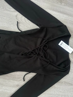 Svart tröja med snörning Gina tricot  - Snygg svart tröja från Gina tricot med lång ärm och ribbad struktur. Framtill har den en djup v-ringning med cool snörningsdetalj som ger en edgy vibe. Perfekt att styla med jeans eller kjol för en trendig look.