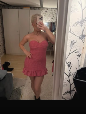 Rosa axelbandslös kort klänning - Säljer en söt rosa axelbandslös kort klänning med volang nertill. Klänningen har en figurnära passform och en strukturerad yta som ger en trendig look. Perfekt för dig som vill sticka ut med en färgglad och feminin stil.
