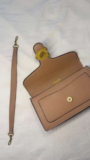 Beige handväska från Coach - Snygg beige handväska från Coach i mjukt läder med avtagbar axelrem. Väskan har guldfärgade metalldetaljer och ett stort C-spänne framtill. Insidan har ett fack med tryckknapp och plats för småsaker. Perfekt för dig som gillar stilrena accessoarer. Pris kan diskuteras 