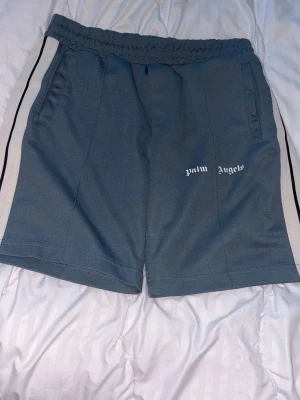 Palm Angels shorts  - Två par Palm Angels shorts i svart och blått med vita sidoränder och logga framtill. De har elastisk midja, sidofickor och sportig look. Perfekta för en avslappnad stil och tillverkade i ett syntetmaterial som ger skön passform.
