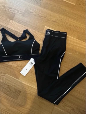 Svart träningsset från Alo Yoga - Snyggt svart träningsset från Alo Yoga med sport-bh och tights. Setet har vita kontrastsömmar och logga på både bh och byxa. Materialet är stretchigt och passar perfekt för gym eller yoga. Sport-bhn har korsade axelband för extra stöd.