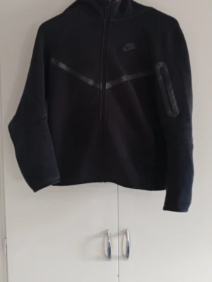 Nike kofta  - Säljer en svart kofta från Nike med hel dragkedja framtill och huva. Hoodien har diskret Nike-logga på bröstet och en snygg detalj med en diagonal linje över bröstet. Materialet ser ut att vara fleece, vilket gör den extra mjuk och varm. Perfekt för chill eller träning.