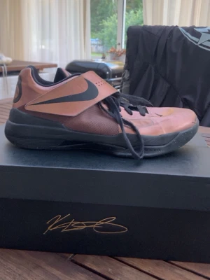 Bronsfärgade Nike sneakers med kardborre - Snygga sneakers från Nike i bronsfärg med svart sula och detaljer. Skon har en låg profil, svart snörning och en bred kardborre över vristen. Materialet är en mix av syntet och mesh för bra andningsförmåga. Perfekta för dig som gillar sportig och unik stil.