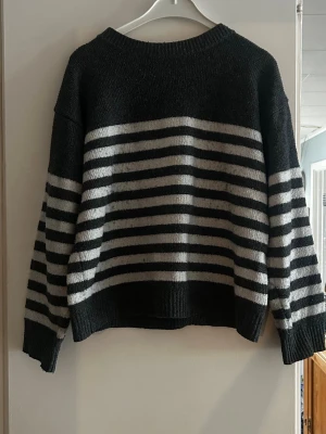 Svartvit randig stickad tröja - Mysig stickad tröja i svart med vita horisontella ränder. Tröjan har rund halsringning, långa ärmar och ribbstickade muddar vid ärmslut och nederkant. Perfekt för dig som gillar en enkel men stilren look.