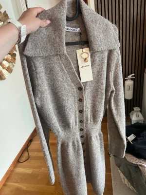Beige stickad långklänning Adoore - Mysig beige stickad långklänning från Adoore med stora slag, markerad midja och knappar framtill. Långärmad och perfekt för kyligare dagar. Enkel men elegant design som passar till många tillfällen. Storlek 32 men passar 34 är väldigt stretchig 