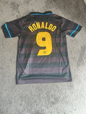Inter Milan Umbro retro matchtröja - Retro Inter Milan matchtröja från Umbro med svart och mörkgråa ränder, gula detaljer och tryck. PIRELLI-logga på bröstet, nummer 9 och RONALDO på ryggen. Krage och ärmslut har blå detaljer. Perfekt för fotbollsfans och samlare. Köptes i retro butik utomlands men var lite för stor