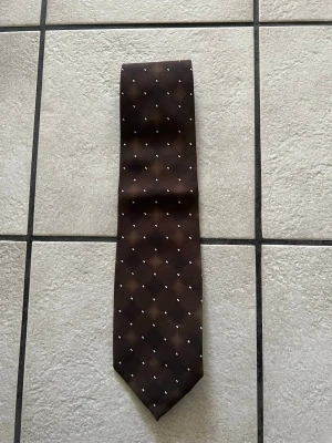 Brun slips med vita prickar JEGEME - Snygg brun slips från JEGEME med små vita prickar i ett rutmönster. Tillverkad i glansigt material som ger en stilren look. Perfekt för att lyfta din outfit med en klassisk accessoar.