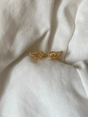 Guldfärgad ring med vingar och stenar - Unik guldfärgad ring med två öppna vingar dekorerade med små gnistrande stenar. Ringen har en öppen design och vingarna är täckta av klara stenar som ger ett lyxigt intryck. Perfekt för dig som gillar detaljer och vill sticka ut.
