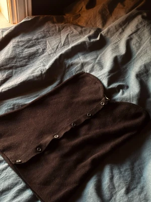 Svart stickad halsduk med knappar - En svart stickad halsduk med knappar längs ena sidan. Halsduken har en enkel och stilren design som gör den lätt att matcha med olika jackor och tröjor. Perfekt för dig som gillar accessoarer med en twist.                                                                              Ser lite brun ut på kameran men är helt klart svart!