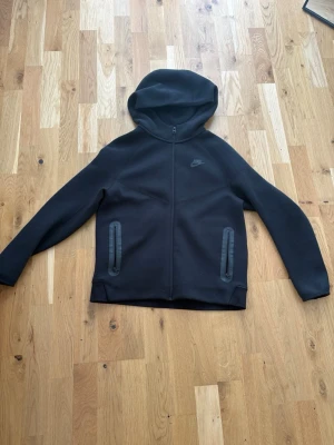svart nike tech - Säljer en svart hoodie från Nike med hel dragkedja framtill och två fickor med dragkedja. Hoodien har en klassisk huva och diskret Nike-logga på bröstet. Materialet är mjukt och stretchigt, perfekt för en avslappnad stil.