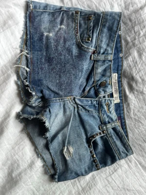 Blå jeansshorts med nitar - Säljer ett par blå denimshorts med råa kanter, slitningar och coola nitar vid fickorna. Shortsen har fem fickor, knappgylf och en avslappnad passform. Perfekta för sommaren och ger en edgy streetstyle-vibe. 