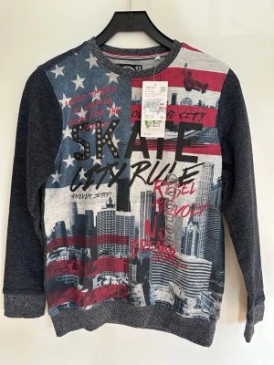 Mörkblå sweatshirt med USA-tryck - Ny oanvänd med tags kvar. Cool sweatshirt med amerikansk flagga, stadssilhuett och text som 'SKATE' och 'CITY RULE'. Tröjan har mörkblå ärmar och baksida, rund hals och ribbade muddar. Perfekt för dig som gillar streetstyle och grafiska tryck. Ekologisk bomull. 
