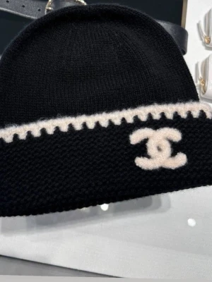 Svart mössa med vit Chanel-logga - Snygg svart stickad mössa från Chanel med vit kant och den klassiska Chanel-loggan broderad framtill. Mössan har en enkel, rundad form och är tillverkad i ett mjukt, värmande material. Perfekt accessoar för att lyfta din outfit.