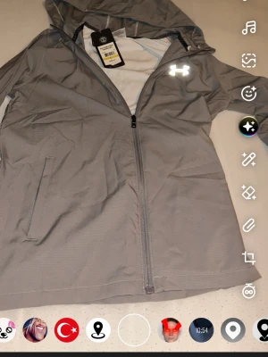 Grå vindjacka från Under Armour - Snygg grå vindjacka från Under Armour med huva och dragkedja framtill. Jackan har långa ärmar, två sidofickor och en stor logga på ryggen. Perfekt för blåsiga dagar och sportiga outfits.