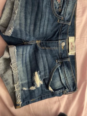 Blå jeansshorts från Hollister - Blå jeansshorts från Hollister med slitna detaljer och råa kanter. Klassisk femficksmodell med knapp och dragkedja framtill. Gula kontrastsömmar och bakfickor med dekorativ söm. Med midjemått på 79cm
