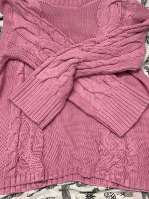 Rosa kabelstickad tröja - Mysig rosa kabelstickad tröja med rund halsringning och breda ribbade muddar. Tröjan har ett klassiskt flätmönster längs ärmar och framstycke, vilket ger en trendig och avslappnad look. Perfekt för lager på lager under kyliga dagar.