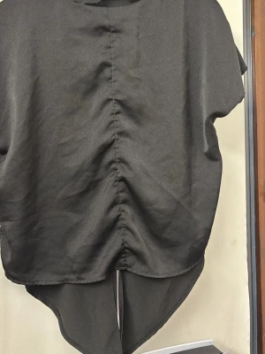 Svart topp med snörning och asymmetrisk fåll - Cool svart topp med korta ärmar och unik asymmetrisk fåll. Toppen har snörning baktill som ger en edgy look och extra detalj. Perfekt för dig som vill sticka ut med en modern och trendig stil. Materialet känns mjukt och lätt mot huden. Den knyts bakifrån och ser finare ut än på bilden.