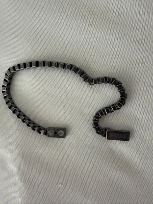 Svart metallarmband från BOSS - Snyggt svart armband från BOSS i metall med fyrkantiga länkar och magnetlås. Armbandet har en modern och stilren design med en diskret logotyp på låset. Perfekt accessoar för dig som gillar minimalistisk och trendig stil.