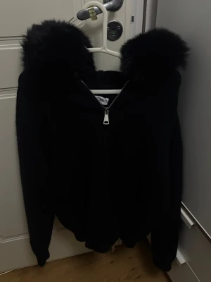Svart hoodie med fuskpäls på huvan - Säljer en svart hoodie med dragkedja och stor huva dekorerad med fluffig fuskpäls. Passar både som en kofta eller som en jacka och är perfekt för alla årstider. Den är i one size och från design by Amor. Endast använd en gång och har inga deffekter. 