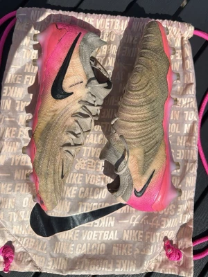 Nike Phantom GT2 Elite fotbollsskor - Säljer ett par Nike Phantom GT2 Elite fotbollsskor i beige och rosa med svarta detaljer. Skorna har en modern, strömlinjeformad design med struktur på ovandelen för bättre bollkontroll. Lätta material och låg profil ger grym känsla på planen. Skorna levereras med en matchande Nike-påse. Skosnören har skador som man kan se på sista bilden och skorna är väl använda och smutsiga men de går att rädda upp. Hör av er vid frågor eller prisförslag😃✅⚽️