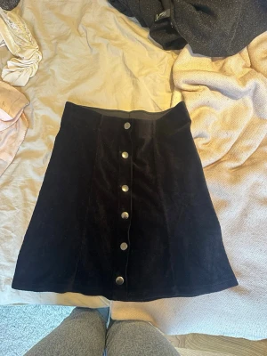 Svart manchesterkjol med knappar - Säljer en svart kortkjol i manchester med snygga silverfärgade knappar framtill. Kjolen har A-linjeform och är perfekt för dig som gillar en klassisk men trendig stil. Passar till många olika outfits och är enkel att matcha.
