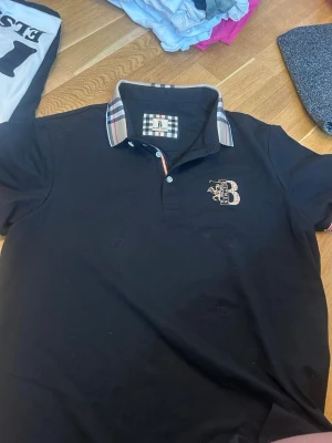 Svart piké med rutiga detaljer Burberry - Svart pikétröja från Burberry med klassisk rutig krage och matchande detalj på bröstet. Tröjan har knappar vid halsen och är kortärmad. Perfekt för en stilren och trendig look.