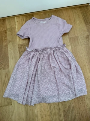 Ljuslila klänning från Mywear Young - Supersöt ljuslila klänning från Mywear Young med ribbad överdel och korta ärmar. Kjolen är mönstrad med små blommor och har en mjuk, luftig känsla. Perfekt för vår och sommar med sin bekväma passform och fina detaljer.