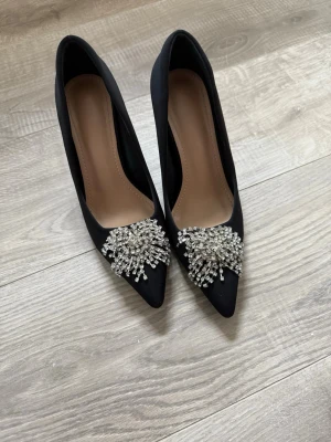 Svarta pumps med pärldetaljer - Stilrena svarta pumps med spetsig tå och dekorativa pärlor framtill. Skorna har en elegant siluett och är tillverkade i ett slätt material som ger en lyxig känsla. Perfekta för dig som vill sticka ut med snygga detaljer.