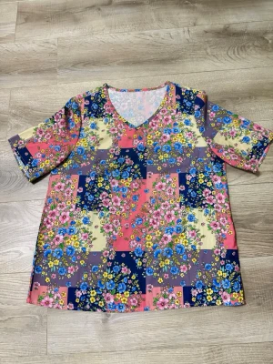 Blommig v-ringad topp i färgglada toner - Färgglad topp med v-ringning och korta ärmar. Mönstret består av blommor i rosa, blått, gult och lila på en mixad bakgrund. Perfekt för dig som gillar färg och mönster. Skön passform och lätt material.