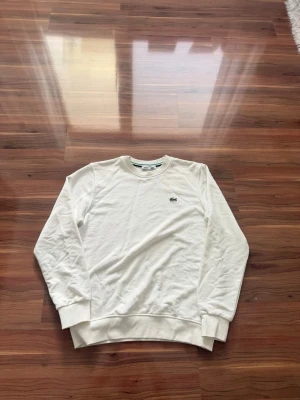 Vit sweatshirt från Lacoste - Klassisk vit sweatshirt från Lacoste med rund halsringning och det ikoniska gröna krokodilmärket broderat på bröstet. Tröjan har ribbade muddar vid ärmslut och nederkant. Perfekt för en clean och stilren look.