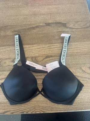 Svart push-up bh Victoria's Secret - Snygg svart push-up bh från Victoria's Secret med vadderade kupor och breda axelband med logotyp. Axelbanden är justerbara och bh:n har hakknäppning bak.