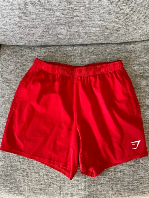 Röda träningsshorts från Gymshark - Snygga röda träningsshorts från Gymshark med elastisk midja och kort passform. Perfekta för gymmet eller löprundan. Tillverkade i ett lätt och snabbtorkande syntetmaterial. Gymshark-logga i vitt på benet för en sportig look.