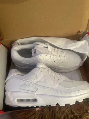 Nike Air Max 90 helvita sneakers - Nike Air Max 90 sneakers i helvitt med klassisk chunky sula och synlig Air-enhet. Ovandelen är i mesh och läder för både stil och komfort. Swoosh-logga på sidan och detaljerade sömmar ger en clean och modern look. Perfekta för dig som gillar streetwear och sportig stil.