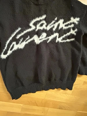 Svart Saint Laurent stickad tröja - Cool svart stickad tröja från Saint Laurent med stor vit logga i borstigt garn framtill. Tröjan har långa ärmar, ribbade muddar och en avslappnad passform. Perfekt för dig som gillar streetstyle och exklusiva märken.