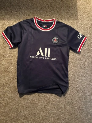 PSG matchtröja 21/22 Mbappé nr 7 - Mörkblå PSG matchtröja storlek 128cm med vita och röda detaljer vid krage och ärmslut. Framsidan har klubbmärke och sponsorlogga, baksidan har Mbappé och nummer 7 tryckt. 