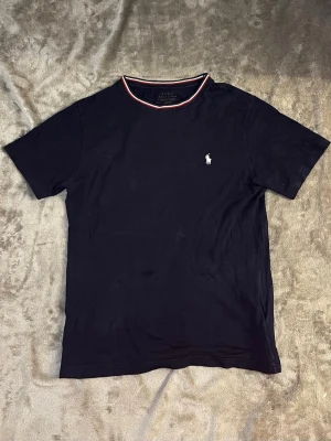 Mörkblå t-shirt från Polo Ralph Lauren - Klassisk mörkblå t-shirt från Polo Ralph Lauren med korta ärmar och rund halsringning med röd och vit kant. Ikoniska broderade loggan i vitt på bröstet. Tillverkad i mjuk bomull för en skön känsla. Och en grisch look. Vid frågor hör av er priset kan alltid diskuteras 