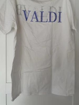 Vit t-shirt från Bvaldi  med strasstenar - Vit t-shirt från Bvaldi med korta ärmar och stora bokstäver i blå och silverfärgade strasstenar på ryggen samt mindre logga framtill. Klassisk rak passform och rund halsringning. Perfekt för dig som gillar lite extra bling i vardagen.