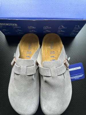 Grå mockatofflor från Birkenstock - Snygga grå tofflor från Birkenstock i mjuk mocka med klassisk slip-in design och justerbart spänne i metall. Fotbädden är i kork och innersulan är bekvämt fodrad. Perfekta för dig som gillar stilrena och bekväma skor.