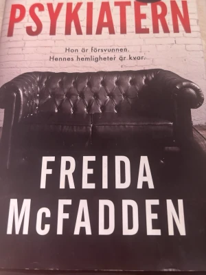 Psykiatern av Freida McFadden - En spännande psykologisk thriller med titeln 'Psykiatern' av Freida McFadden. Omslaget är svartvitt med en bild på en klassisk lädersoffa mot en vit tegelvägg. Perfekt för dig som gillar mysterier och nervkittlande berättelser.