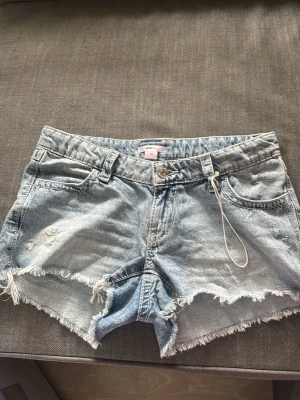 Ljusblå jeansshorts med broderi - Säljer ett par ljusblå jeansshorts från Gina tricot med fransiga kanter och slitningar framtill. Shortsen har broderade fickor bak med coolt mönster och klassisk femficksdesign. Perfekta för sommaren och ger en avslappnad vibe.