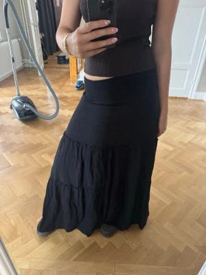 Svart långkjol med volangdetaljer - Säljer en svart långkjol med bred smock i midjan och flera volanglager. Kjolen har en flowy passform och är perfekt för en avslappnad och trendig look. Passar till både linne och stickad tröja.