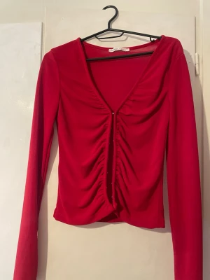 Röd långärmad topp med snörning - Trendig röd topp med lång ärm och snygg v-ringning. Framtill har toppen en rynkad snörningsdetalj som ger en cool look. Materialet känns mjukt och stretchigt, perfekt för att styla med jeans eller kjol. Inga synliga loggor eller märken.