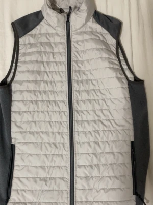 Jack Jones väst  - Snygg ljusgrå dunväst med mörkgrå sidor och axelparti. Västen har quiltad design, hel dragkedja framtill och två fickor med dragkedja. Perfekt för lager-på-lager och håller dig varm utan att bli för tung. Sportig och stilren look.