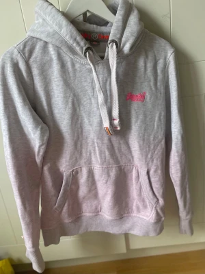 Ljusgrå/rosa hoodie från Superdry - Säljer en ljusgrå/rosa hoodie från Superdry🌸. Storleken är M men om man vill ha lite plats så är det mer som en S. Tillverkad i mjukt bomullsmaterial som är perfekt för chill dagar.