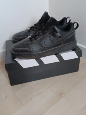 Svarta Nike Air Force 1 sneakers - Klassiska helsvarta Nike Air Force 1 sneakers i läder med lågt skaft och ikonisk Swoosh på sidan. Skorna har svart yttersula, svarta skosnören och diskreta detaljer. Perfekta för en clean och stilren look!