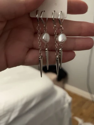 Hemma gjorda Silvriga örhängen med pärlor och spikes - Hemma gjorda Snygga hängande örhängen i silverfärgad metall med vita pärlor och spetsiga spikes. Kedjedetaljer ger en edgy vibe och pärlorna adderar en touch av elegans. Perfekta för dig som gillar att sticka ut med dina accessoarer.
